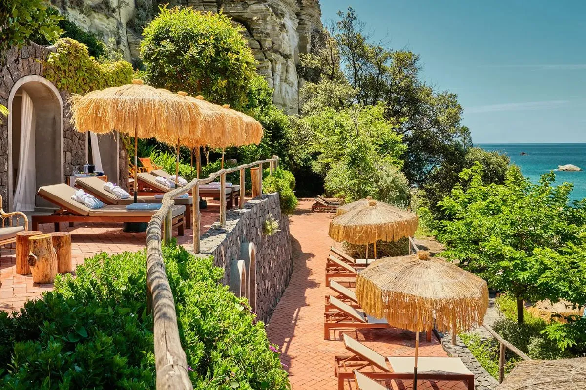 Terrazza panoramica con lettini e ombrelloni di paglia tra la vegetazione e la scogliera sul mare