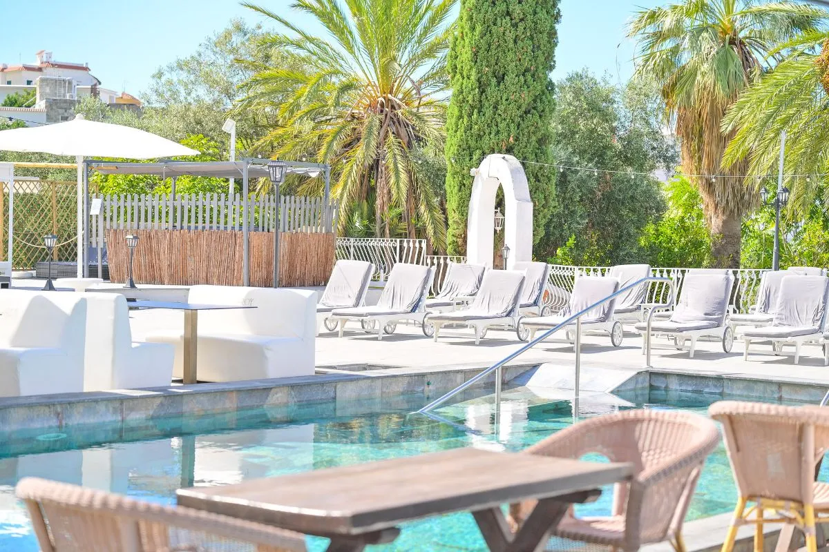 Bordo piscina con lettini, divanetti bianchi e palme sotto cielo azzurro