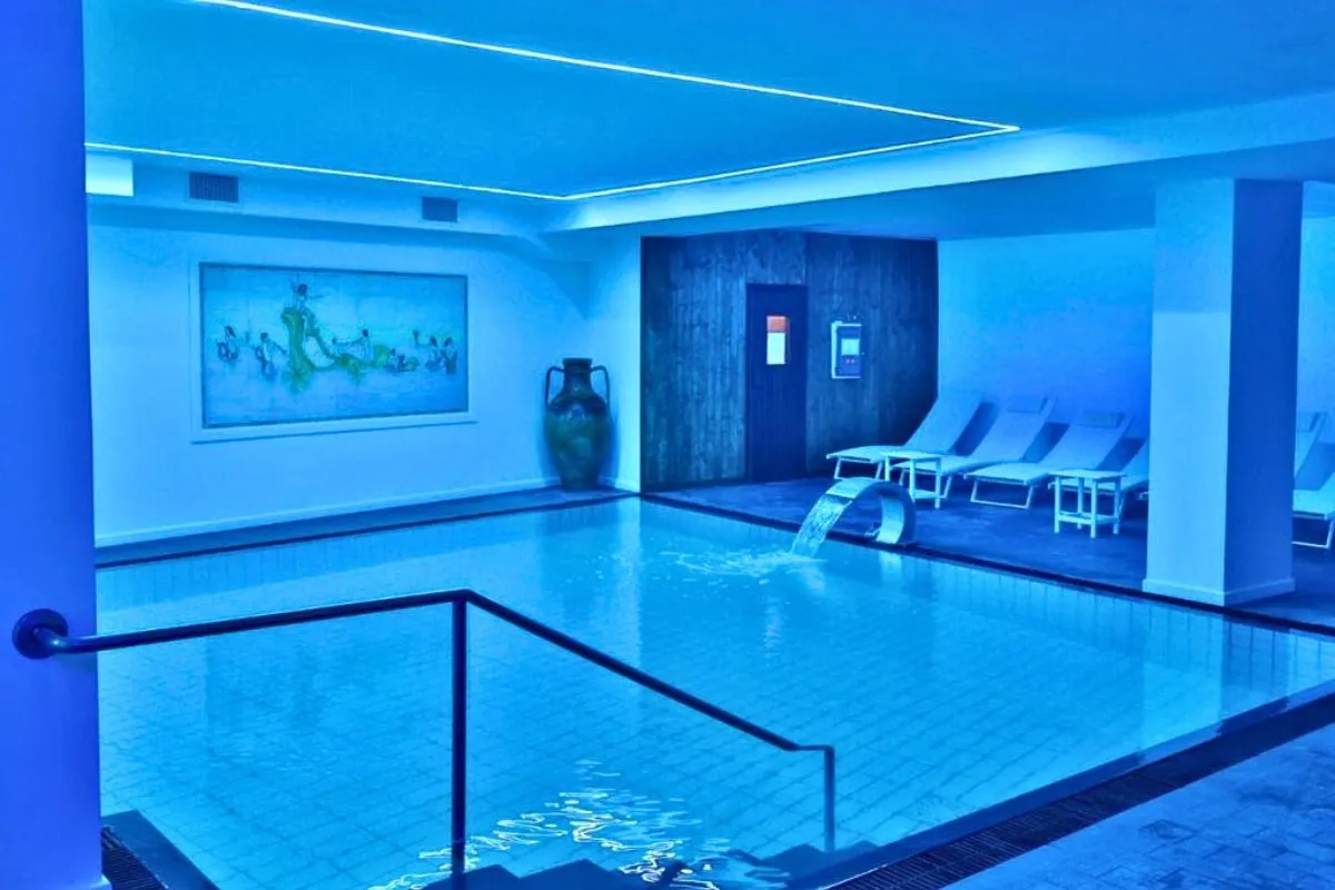 Piscina interna con illuminazione blu, cascata, lettini e sauna in legno