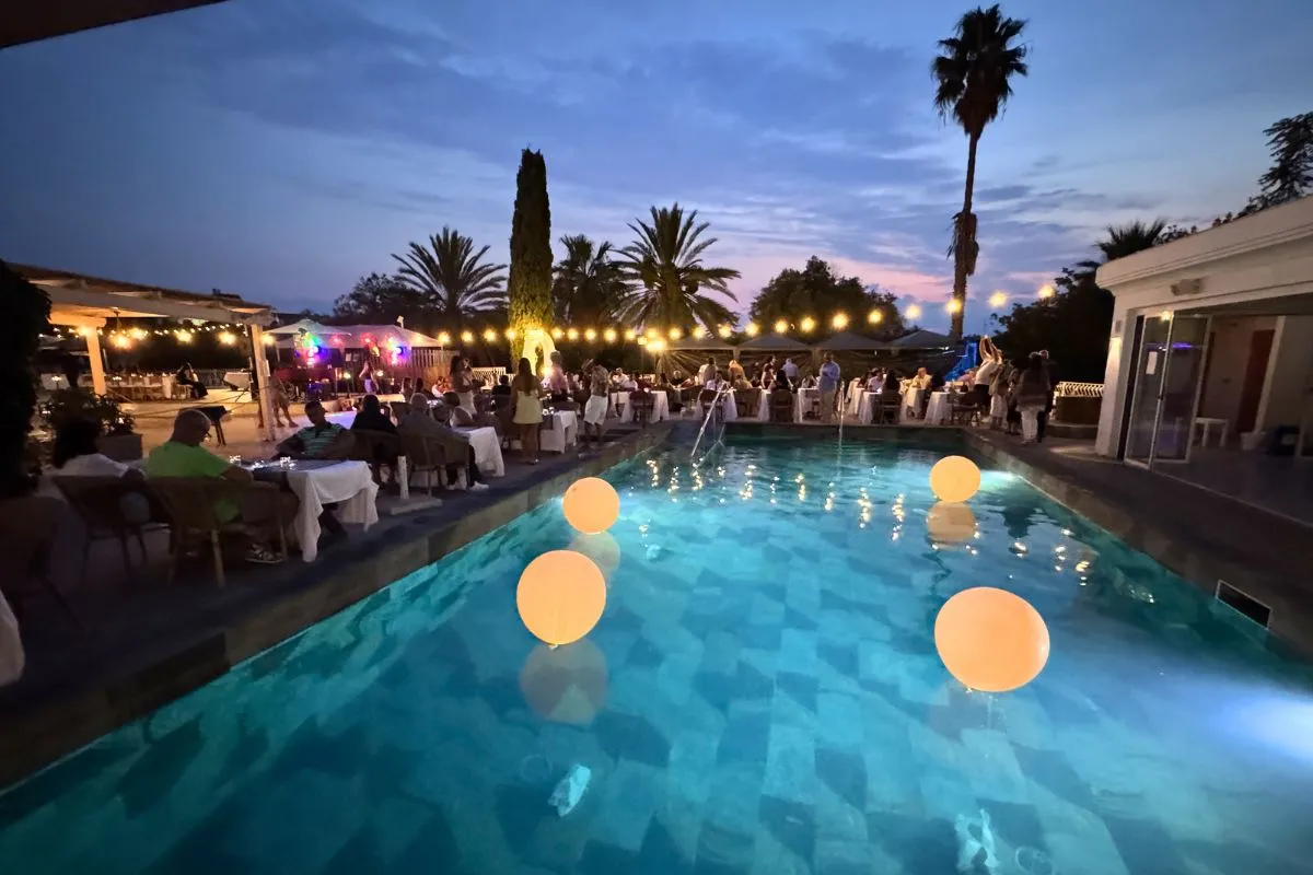 Festa serale a bordo piscina con tavoli apparecchiati, luci e palloncini luminosi in acqua