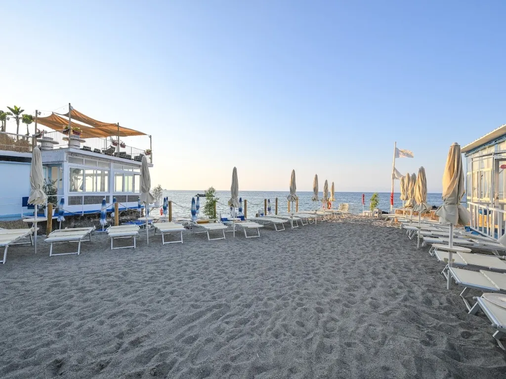 Spiaggia di sabbia scura con lettini, ombrelloni chiusi e stabilimento balneare fronte mare