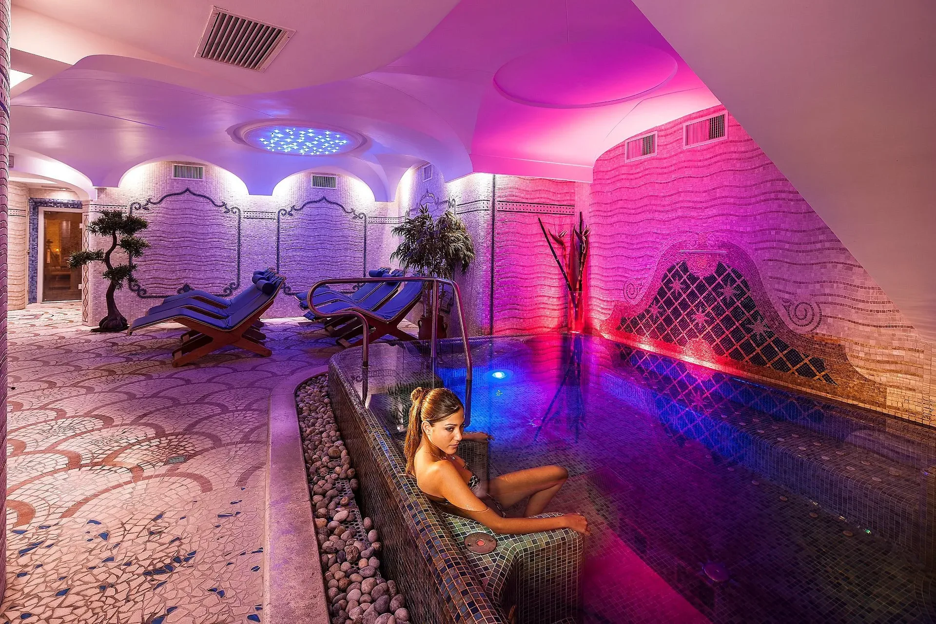 Area spa con illuminazione viola e rosa, lettini relax e vasca idromassaggio a mosaico