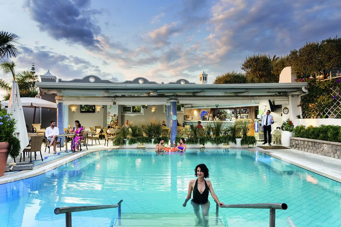 Piscina al tramonto con bar e ospiti immersi nell'acqua nell'atmosfera serale del resort