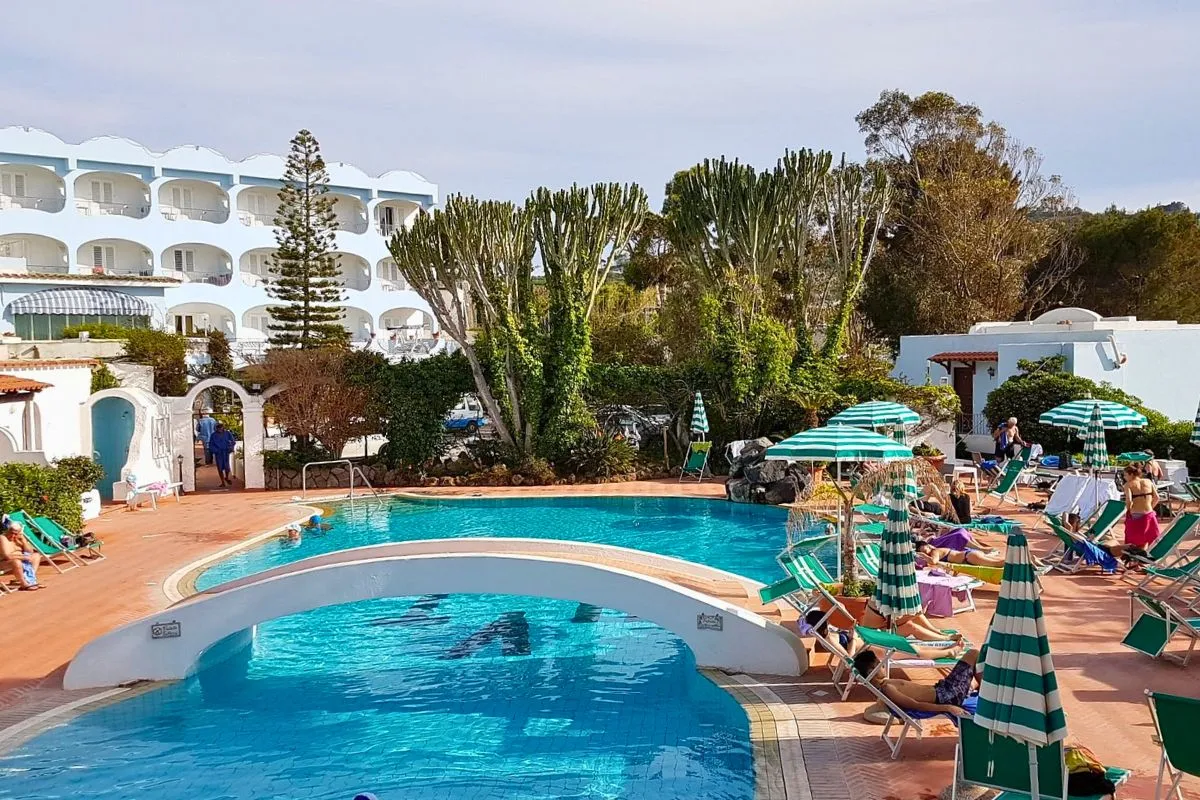 Piscina esterna con ospiti, lettini e ombrelloni a righe verdi davanti all'edificio bianco dell'hotel