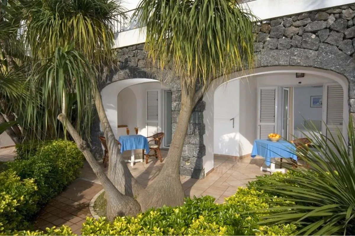 Patio esterno di una camera con muri in pietra lavica, tavolini con tovaglie azzurre e giardino tropicale